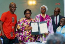 Five professionals pose for a photo as two women hold an open certificate in a formal awards ceremony, flanked by colleagues in colorful attire and a man in a suit on the right side of the group.