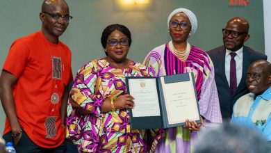 Five professionals pose for a photo as two women hold an open certificate in a formal awards ceremony, flanked by colleagues in colorful attire and a man in a suit on the right side of the group.