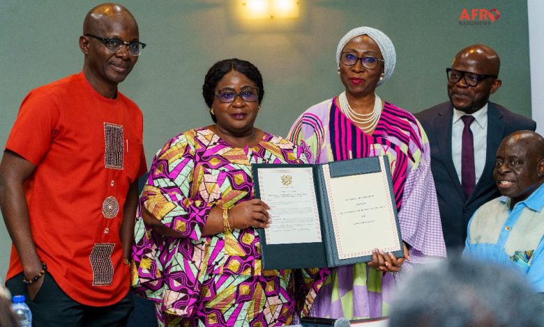 Five professionals pose for a photo as two women hold an open certificate in a formal awards ceremony, flanked by colleagues in colorful attire and a man in a suit on the right side of the group.