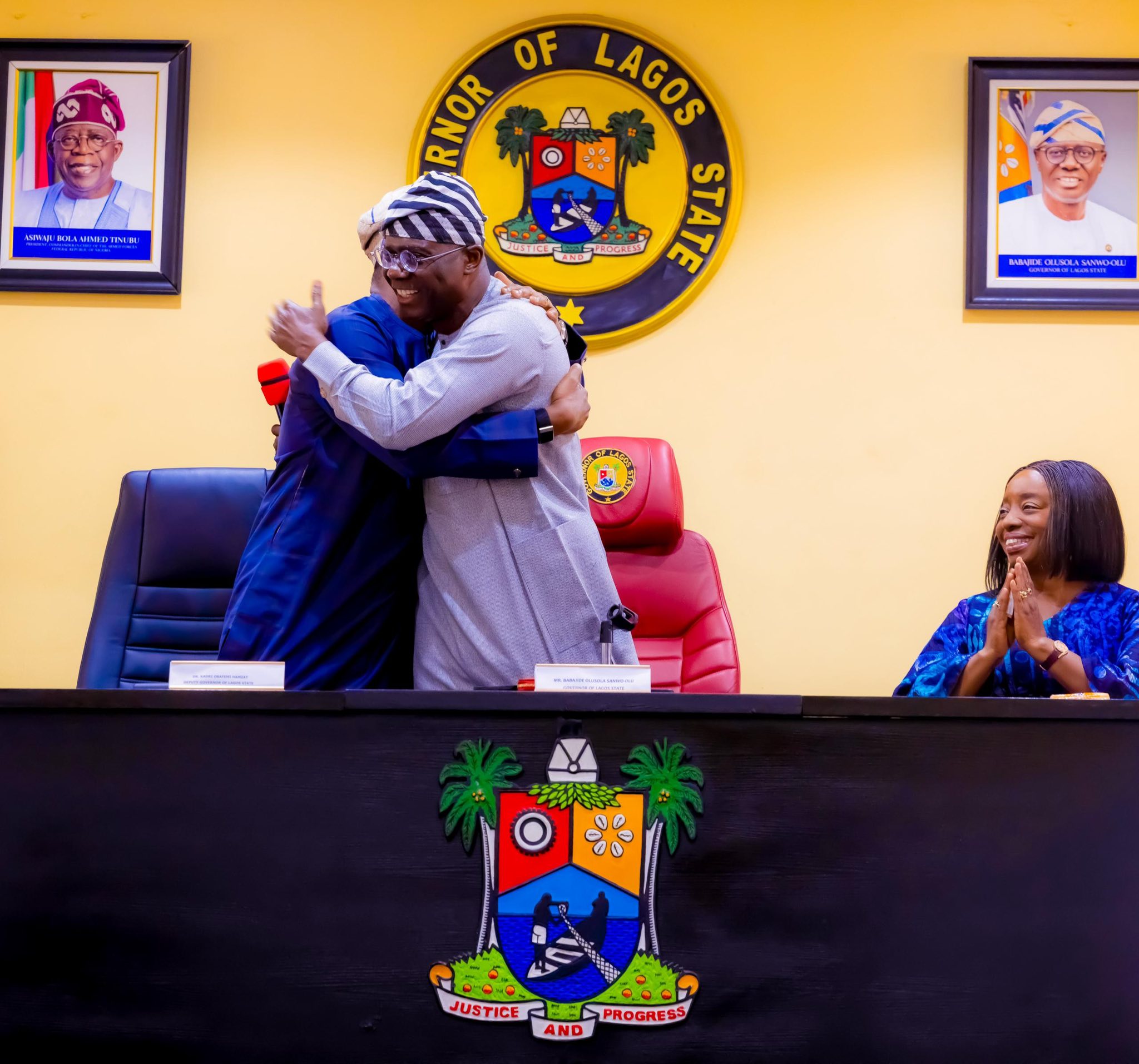 Two men hug in front of Lagos State emblem on a yellow wall, with framed portraits on either side and a smiling woman clapping nearby.