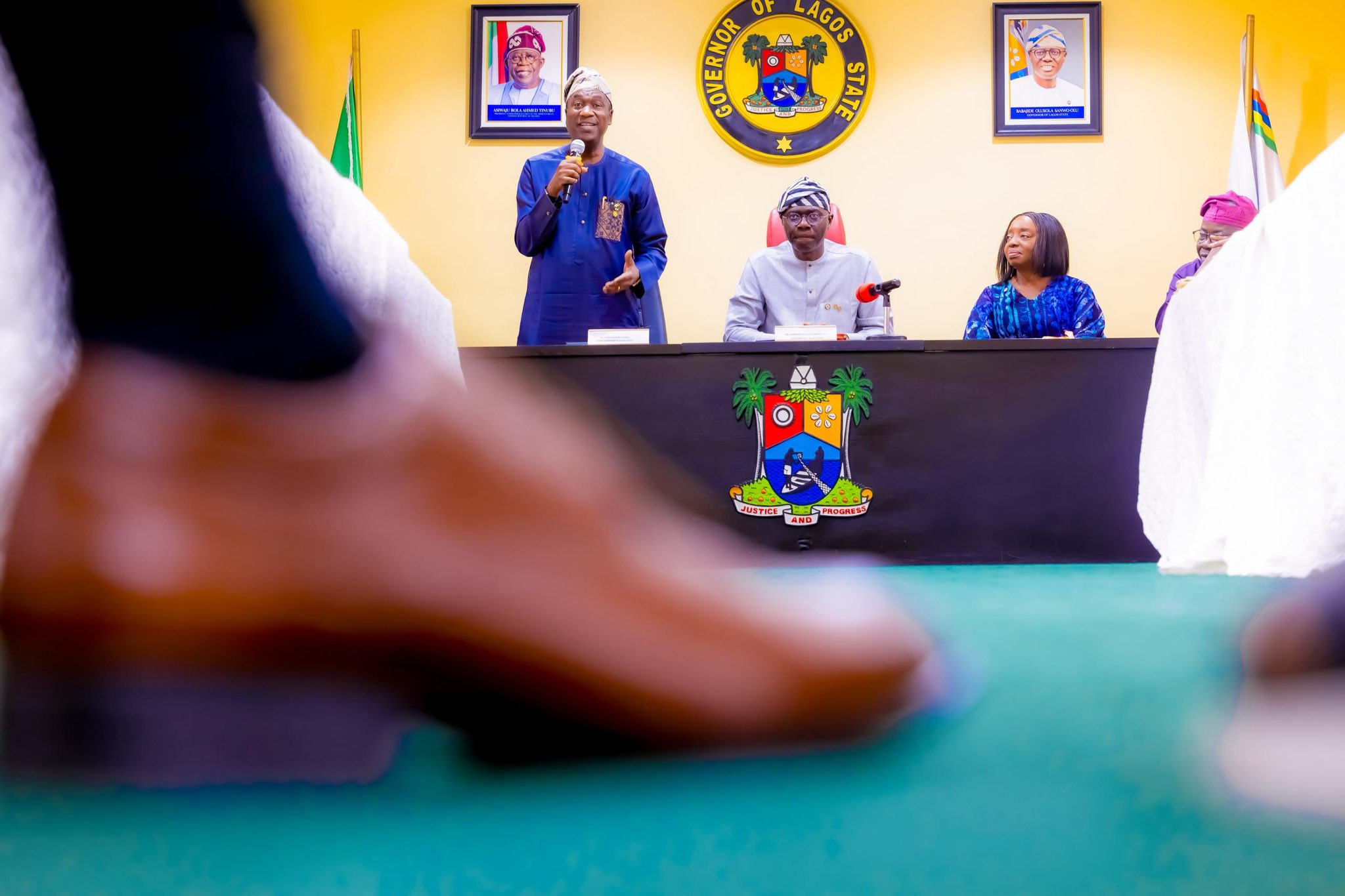 Man in blue traditional attire stands and speaks into a microphone at a government conference desk; Lagos State seal and portraits on the yellow wall behind, others seated nearby.