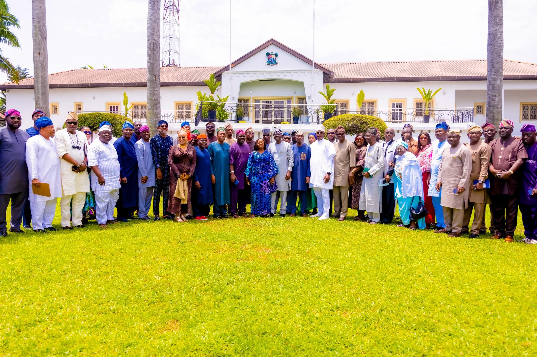 Large group of people in colorful traditional Nigerian attire posing on a lawn in front of a white building with palm trees.