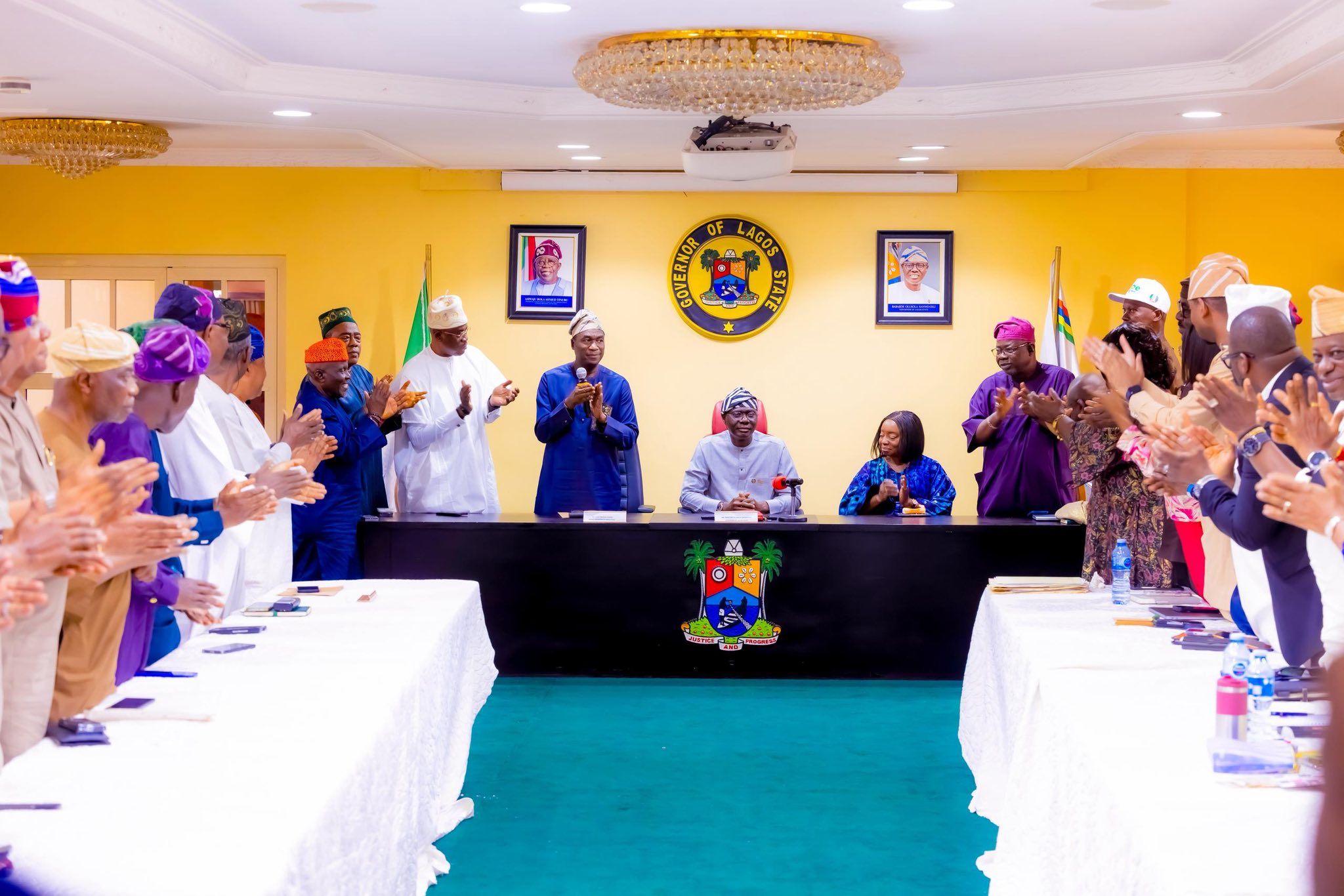 Officials in colorful traditional attire clap around a long conference table as a central speaker addresses the room; Lagos State seal on wall.