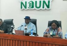 Panel of five people seated at a conference table with microphones; a man in a blue shirt and hat speaks, NDUN branding visible behind them.