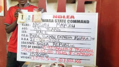 Man in a red shirt stands beside a white information board from NDLEA Warra State Command, showing detainee details (name Nasiru Mazu, age 33, male) and a drug seizure note.