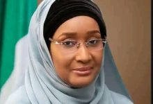 Portrait of a woman wearing a light blue hijab and glasses, looking at the camera with a gentle smile.