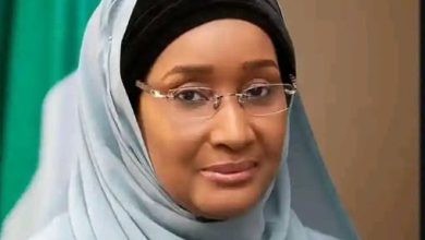 Portrait of a woman wearing a light blue hijab and glasses, looking at the camera with a gentle smile.