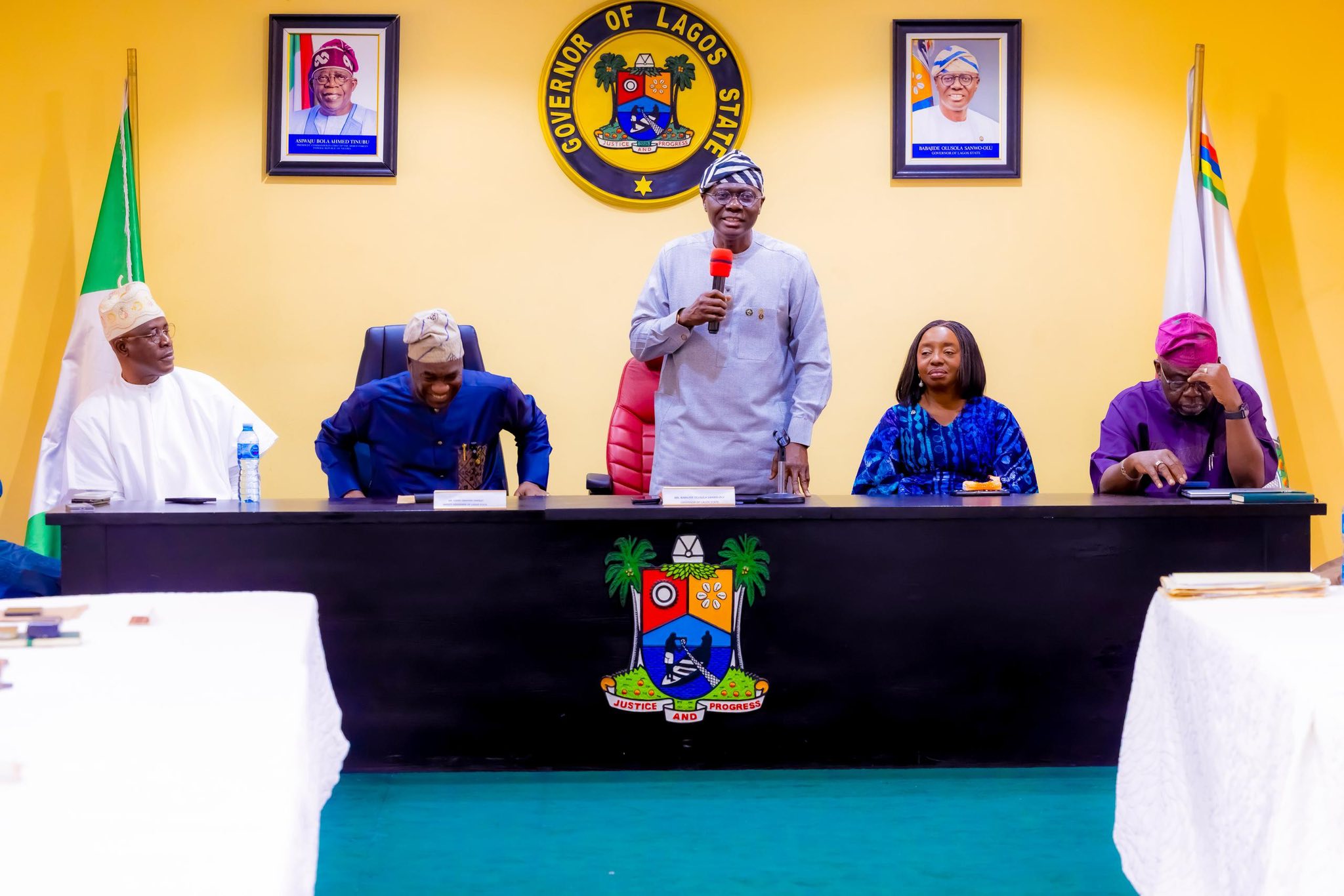 Man stands at a Lagos State government panel, speaking into a red microphone, flanked by four officials at a long table; official seal and flags on yellow wall behind them.