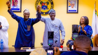 Two men in traditional Nigerian attire raise their clasped hands in celebration at a Lagos State press conference, with the state seal on the wall behind.