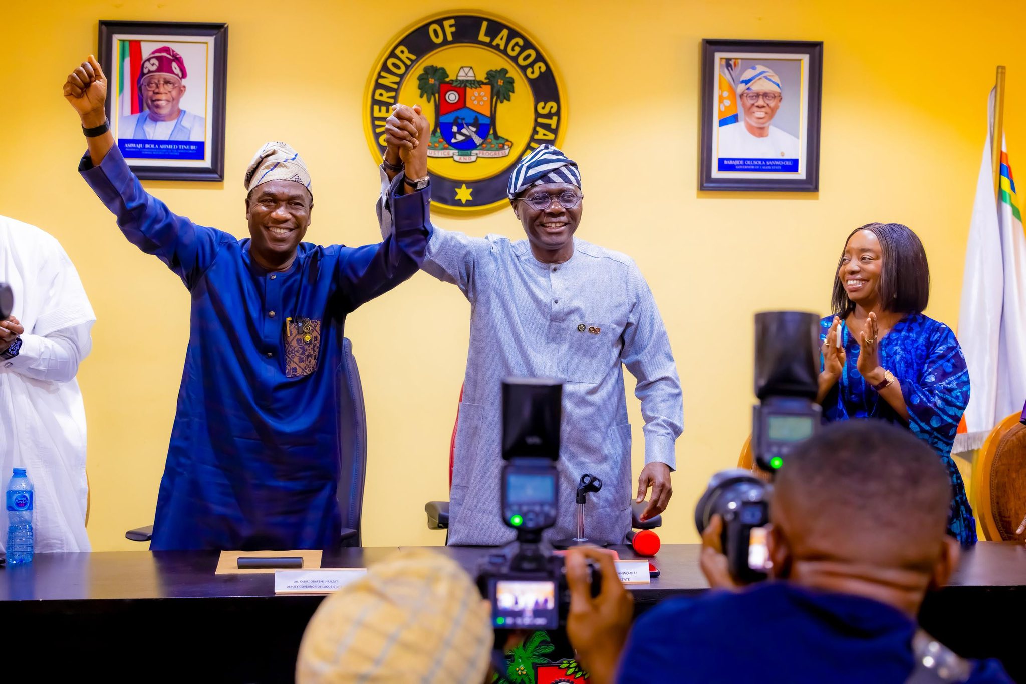 Two men in traditional Nigerian attire raise their clasped hands in celebration at a Lagos State press conference, with the state seal on the wall behind.