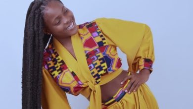Smiling Black woman in a bright yellow wrap outfit with a colorful patterned scarf, standing with hands on hips against a light blue background.
