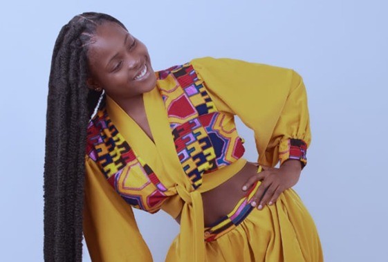 Smiling Black woman in a bright yellow wrap outfit with a colorful patterned scarf, standing with hands on hips against a light blue background.