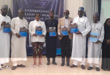 Group of people in traditional and formal attire posing on a stage, each holding a blue booklet, with a dark banner and a projection screen behind them.