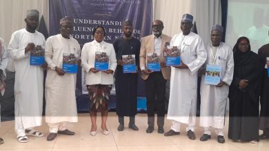 Group of people in traditional and formal attire posing on a stage, each holding a blue booklet, with a dark banner and a projection screen behind them.
