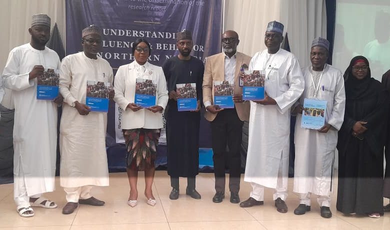 Group of people in traditional and formal attire posing on a stage, each holding a blue booklet, with a dark banner and a projection screen behind them.