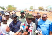 Man in a gray suit and green fedora speaks to reporters holding microphones at a roadside press briefing with onlookers nearby.