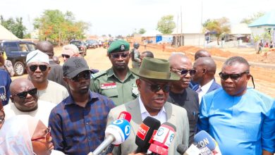 Man in a gray suit and green fedora speaks to reporters holding microphones at a roadside press briefing with onlookers nearby.