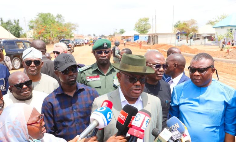 Man in a gray suit and green fedora speaks to reporters holding microphones at a roadside press briefing with onlookers nearby.