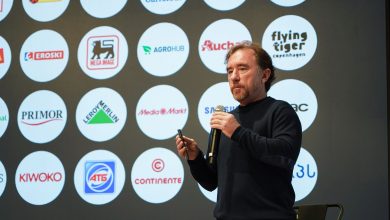 Andrea di Fonzo - The Times of Abuja Male presenter on stage holding a microphone with a wall of circular sponsor logos behind him, facing the audience.