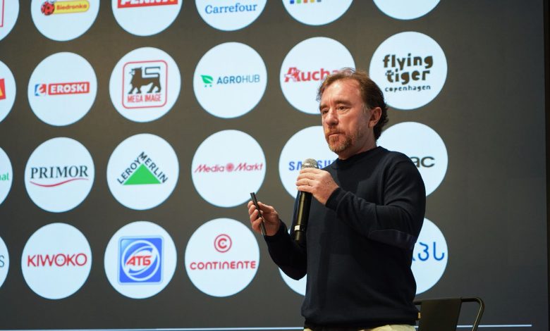 Male presenter on stage holding a microphone with a wall of circular sponsor logos behind him, facing the audience.