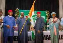 Group of people in colorful traditional Nigerian attire on a stage, a man holds a Nigerian flag while others applaud or speak.