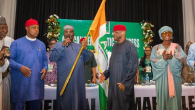 Group of people in colorful traditional Nigerian attire on a stage, a man holds a Nigerian flag while others applaud or speak.