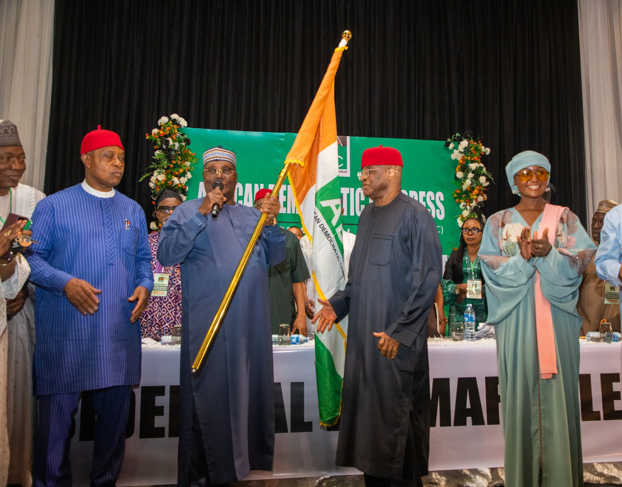 Atiku wins 1 - The Times of Abuja Group of people in colorful traditional Nigerian attire on a stage, a man holds a Nigerian flag while others applaud or speak.