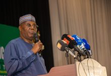 African man in blue traditional outfit and patterned cap speaks into a handheld microphone at a press conference with multiple news microphones at the podium