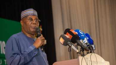 African man in blue traditional outfit and patterned cap speaks into a handheld microphone at a press conference with multiple news microphones at the podium