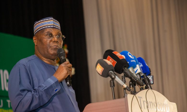 Atiku wins 2 - The Times of Abuja African man in blue traditional outfit and patterned cap speaks into a handheld microphone at a press conference with multiple news microphones at the podium