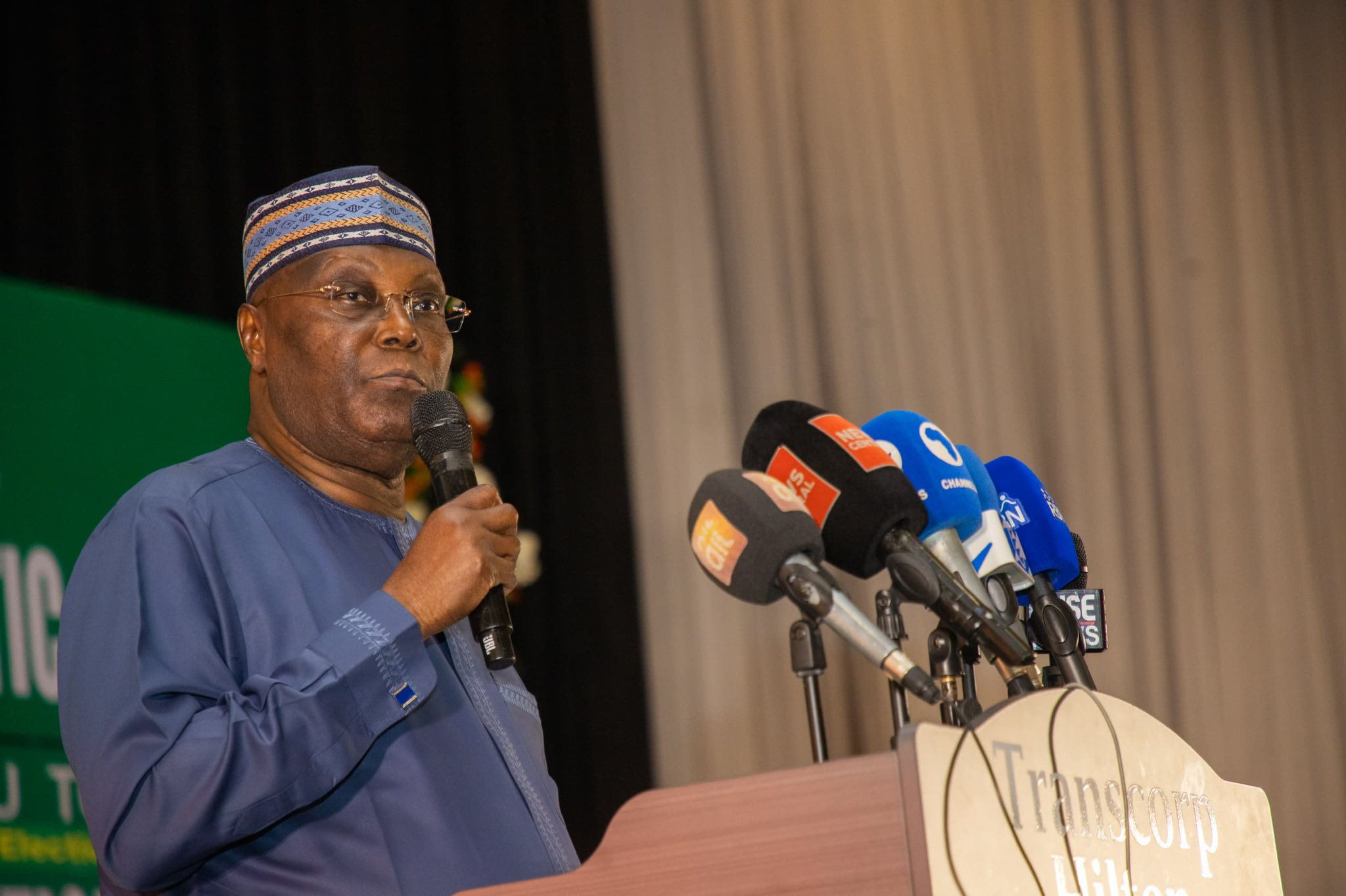 Atiku wins 2 - The Times of Abuja African man in blue traditional outfit and patterned cap speaks into a handheld microphone at a press conference with multiple news microphones at the podium