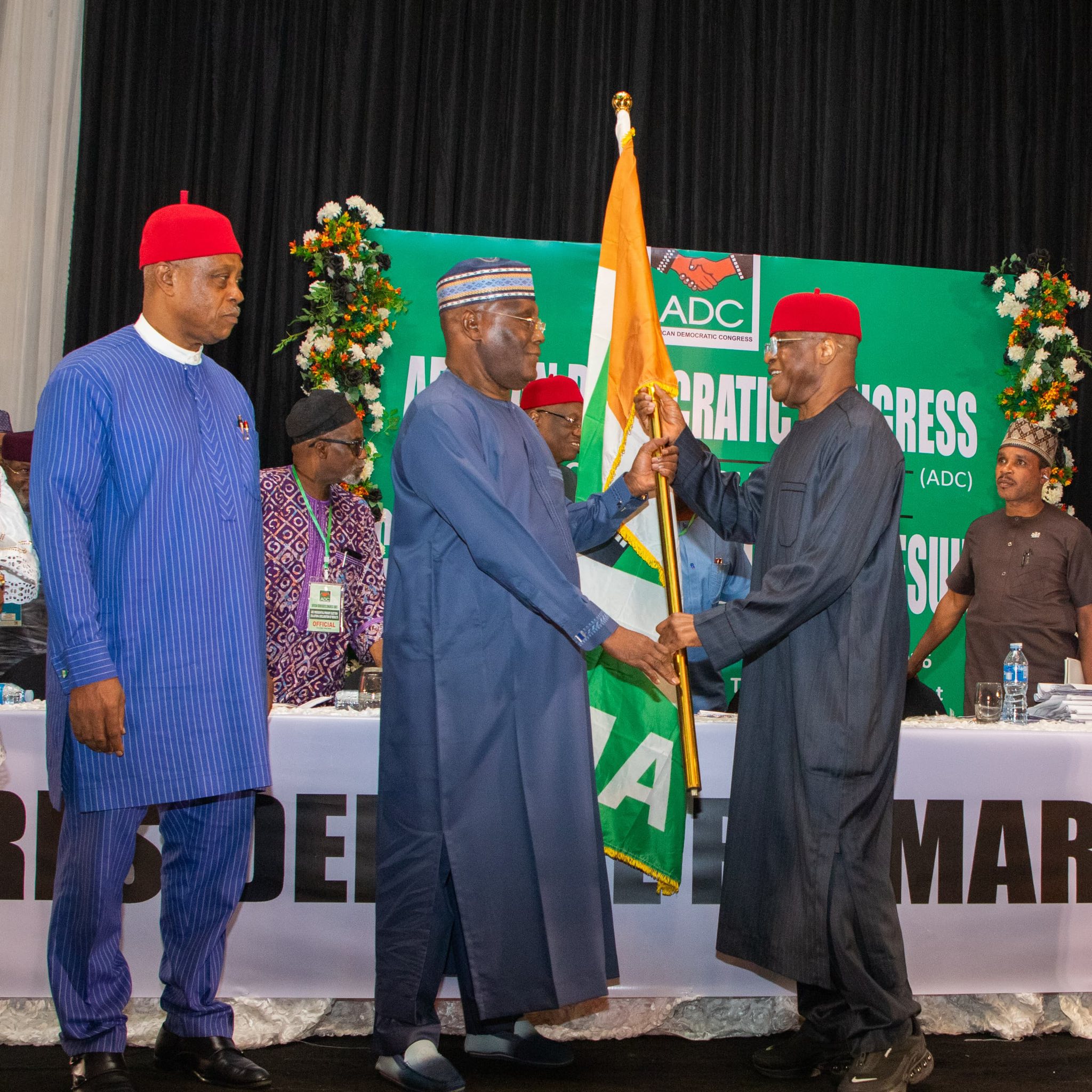 Atiku wins 3 - The Times of Abuja Men in traditional Nigerian attire on stage at a political event, exchanging a flag with a green banner in the background.