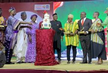 Group of people in colorful traditional attire presenting a tall multi-tiered cake on stage at an awards event.