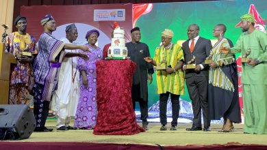 Group of people in colorful traditional attire presenting a tall multi-tiered cake on stage at an awards event.