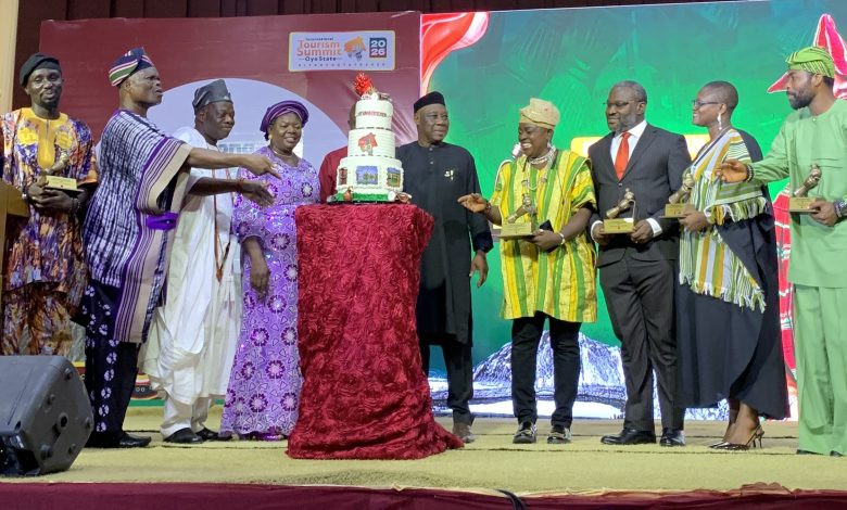 Group of people in colorful traditional attire presenting a tall multi-tiered cake on stage at an awards event.