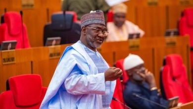 Jibrin Echocho 2 - The Times of Abuja Politician speaking at a microphone in a legislative chamber with red theater-style seats behind him.