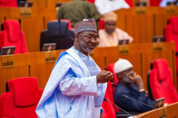 Jibrin Echocho 2 - The Times of Abuja Politician speaking at a microphone in a legislative chamber with red theater-style seats behind him.