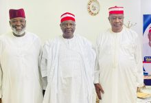 Three men in white traditional outfits with red caps posing indoors, a clock on the wall behind them and a political poster to the right side.