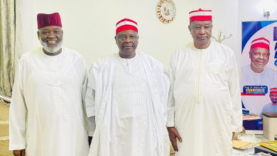 Kwankwaso - The Times of Abuja Three men in white traditional outfits with red caps posing indoors, a clock on the wall behind them and a political poster to the right side.