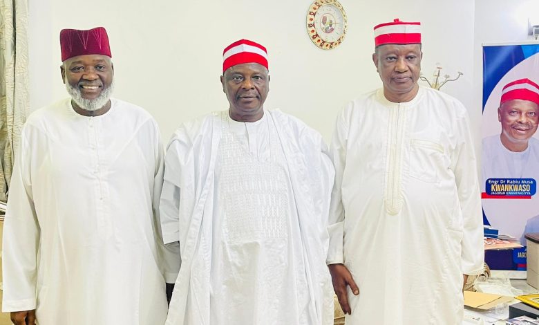 Three men in white traditional outfits with red caps posing indoors, a clock on the wall behind them and a political poster to the right side.
