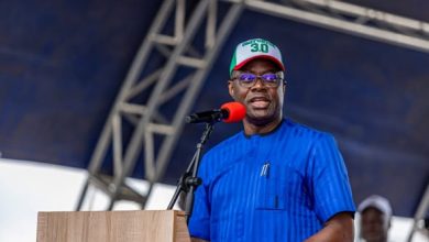 Makinde - The Times of Abuja Man in a blue traditional outfit speaks at a wooden podium on a stage, wearing a white cap with green and red accents and a red microphone in front of him.