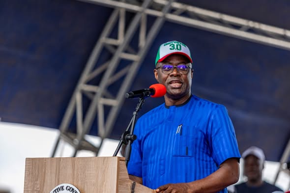 Makinde - The Times of Abuja Man in a blue traditional outfit speaks at a wooden podium on a stage, wearing a white cap with green and red accents and a red microphone in front of him.