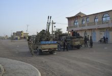 Military convoy with soldiers and a rocket launcher mounted on trucks in a dusty town street.