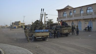 Military convoy with soldiers and a rocket launcher mounted on trucks in a dusty town street.