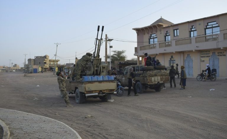 Military convoy with soldiers and a rocket launcher mounted on trucks in a dusty town street.
