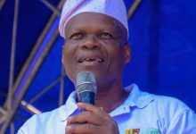 Smiling man wearing a white headwrap speaks into a microphone on a blue stage backdrop.