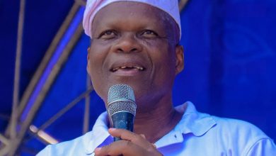 Smiling man wearing a white headwrap speaks into a microphone on a blue stage backdrop.