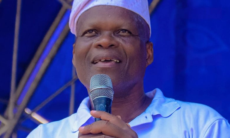 Smiling man wearing a white headwrap speaks into a microphone on a blue stage backdrop.