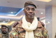 Man in camouflage military uniform and beret, wearing a light scarf, standing in an indoor hallway.”