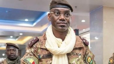 Man in camouflage military uniform and beret, wearing a light scarf, standing in an indoor hallway.”